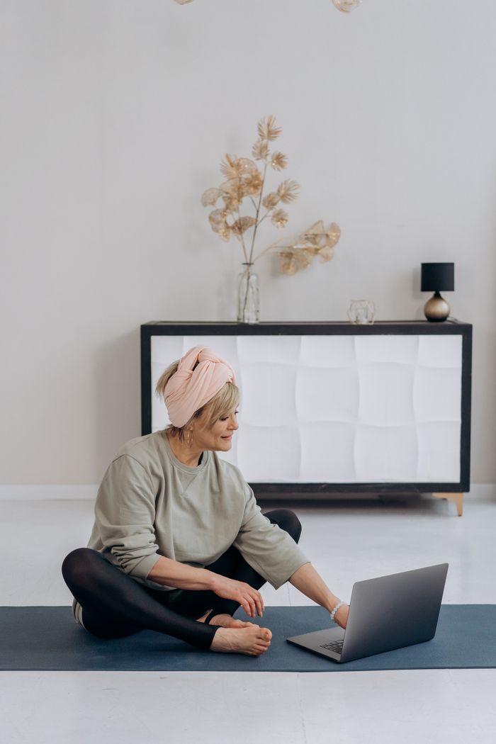 Elderly woman sitting on a yoga mat using a laptop in a stylish living room.